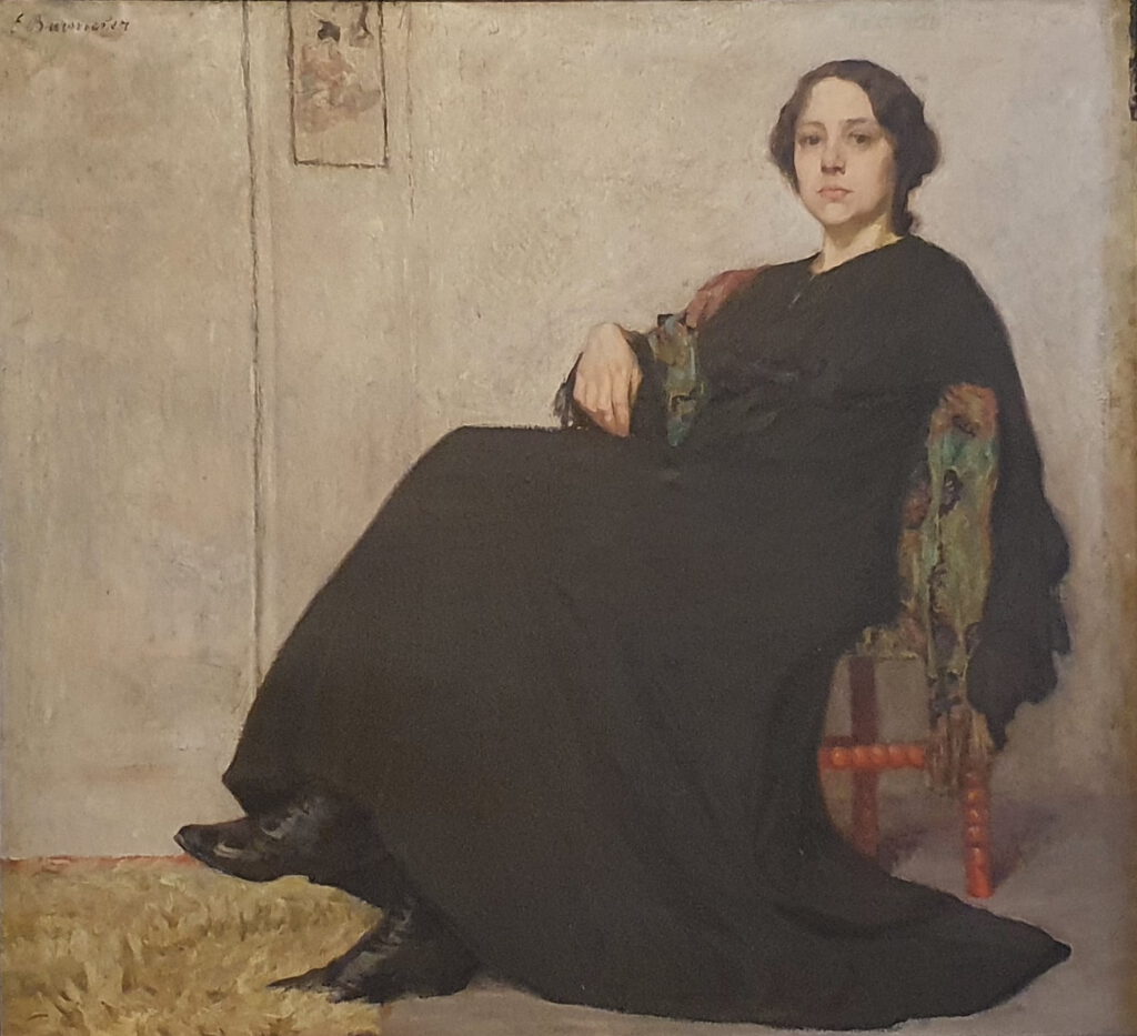 Eine junge Frau mit hochgesteckten Haaren und in einem weiten schwarzen Kleid gemütlich auf einem Stuhl sitzend.