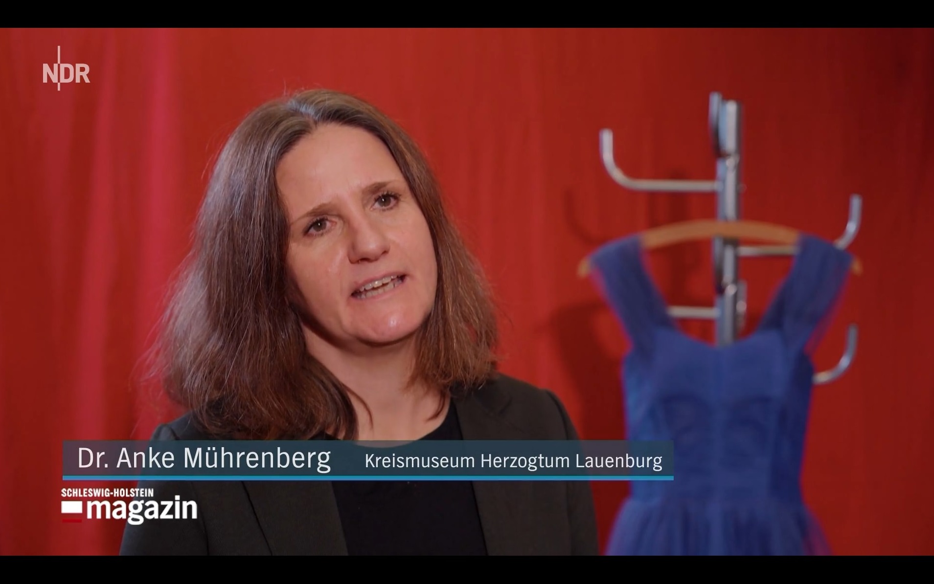 Museumsleiterin Dr. Anke Mührenberg vor rotem Vorhang, neben ihr auf einem Kleiderständer ein blaues Cocktailkleid.